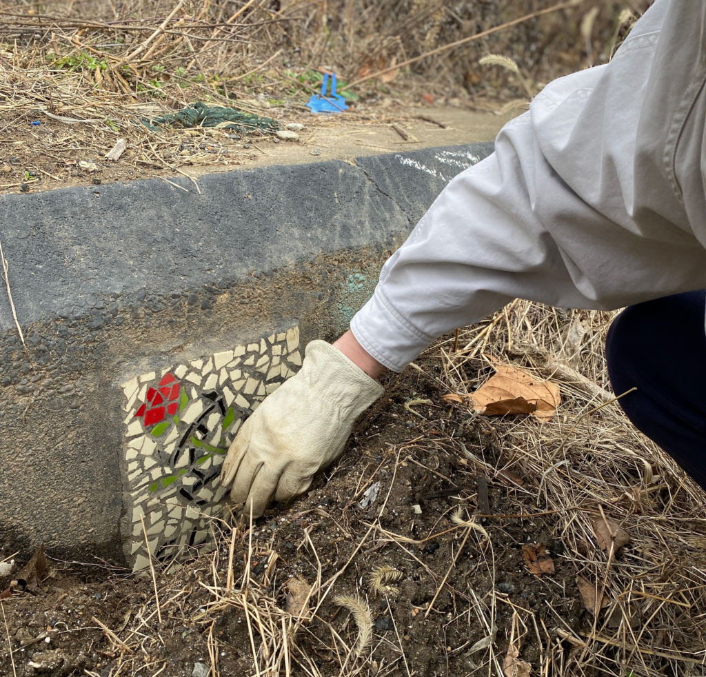 gloved hand brushing dirt off of a mosaic rose embedded in a retaining wall